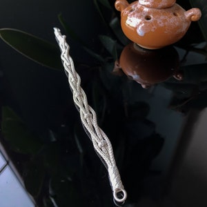May include: A silver wand with a braided design and a loop at the end. Next to it is a small, brown ceramic teapot with a lid and holes. The objects are on a reflective black surface.