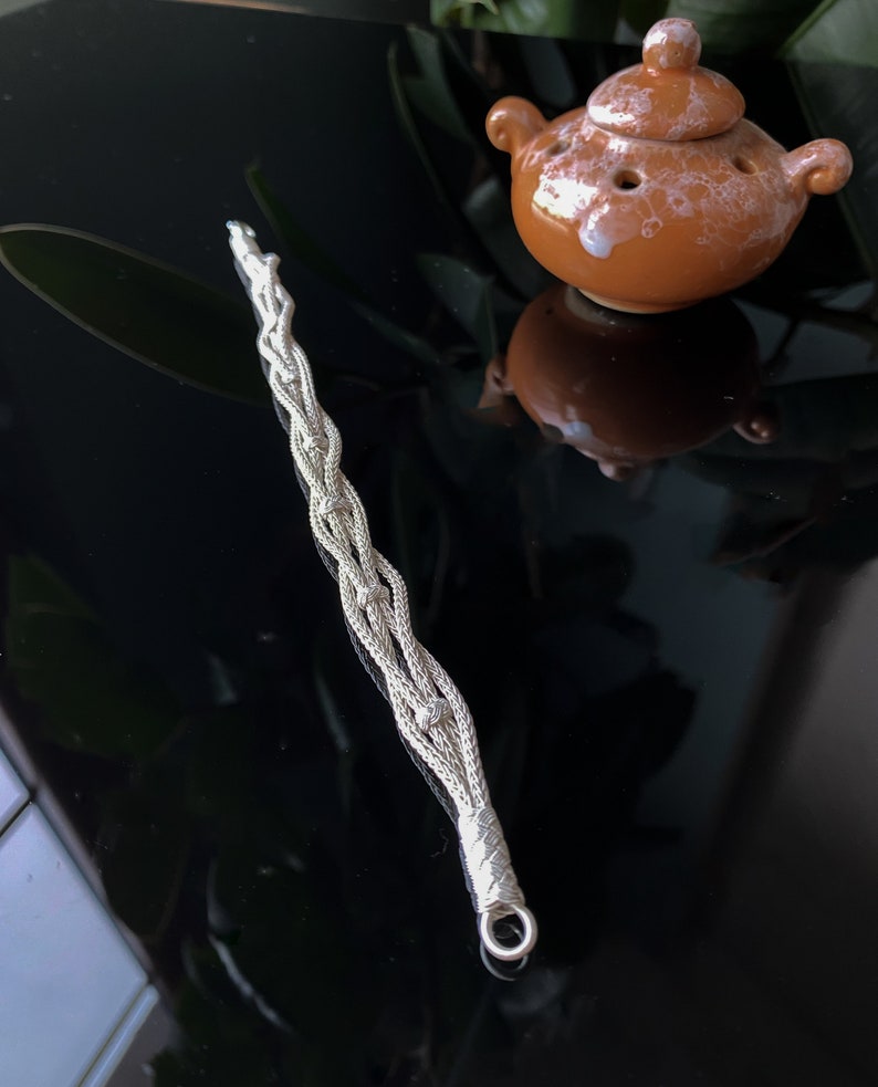May include: A silver wand with a braided design and a loop at the end. Next to it is a small, brown ceramic teapot with a lid and holes. The objects are on a reflective black surface.