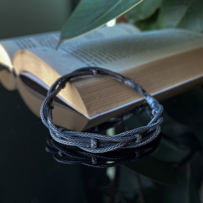 May include: A silver braided bracelet with multiple strands, lying on a reflective surface in front of an open book. The bracelet has a textured appearance with small knots. A green plant is visible in the background.