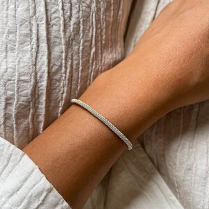 May include: A silver chain bracelet with a delicate, woven design. The bracelet is worn on a person's wrist.