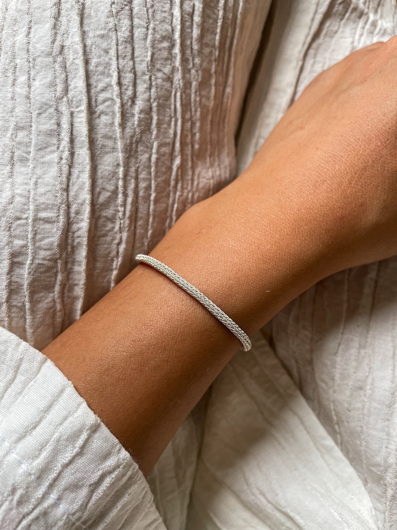 May include: A silver chain bracelet with a delicate, woven design. The bracelet is worn on a person's wrist.