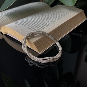 May include: A silver bracelet with a woven design lies on an open book. The bracelet is made of multiple strands of silver, creating a textured appearance. The book's pages are aged, and the background is dark.