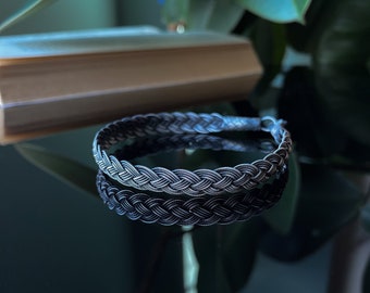 Op de afbeelding: Een zilverkleurige armband met een gevlochten ontwerp. De armband is gemaakt van meerdere strengen die met elkaar zijn verweven, waardoor een textuur ontstaat. De armband wordt gepresenteerd op een reflecterend oppervlak, met een boek op de achtergrond.