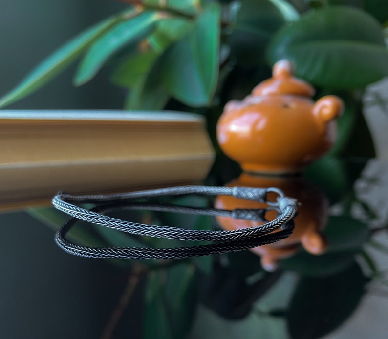 May include: A black braided chain necklace with a silver clasp. The necklace is lying on a reflective surface.