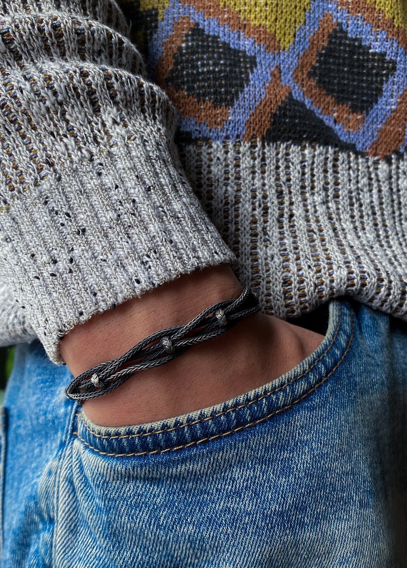 May include: A silver bracelet with a braided design and three decorative knots. The bracelet is worn on a wrist, with a blue jeans pocket in the background. The person is wearing a grey sweater with a patterned design.