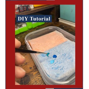 May include: A DIY tutorial for creating a wet palette using a plastic container, paper towels, and paint. The image shows a close-up of the wet palette with a paintbrush and paint drops on the paper towels. The text "DIY Tutorial" is visible in the image.