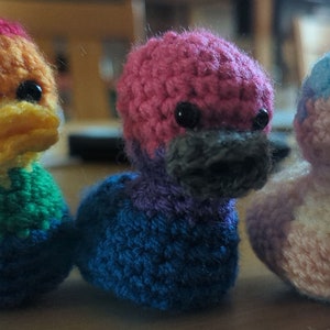 May include: Three crocheted duck plushies, each with a different colour scheme. The first duck is orange, yellow, green, and blue. The second duck is pink, purple, and blue. The third duck is pink, white, and blue.