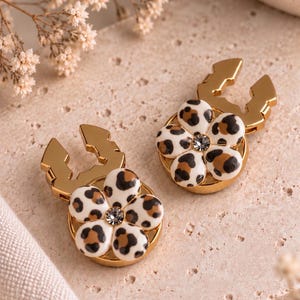 Peut inclure: Une paire de boucles d'oreilles en métal doré avec une base circulaire et une breloque en forme de fleur. Chaque fleur présente des pétales blancs avec un motif léopard marron et noir et un petit cristal transparent et brillant au centre.