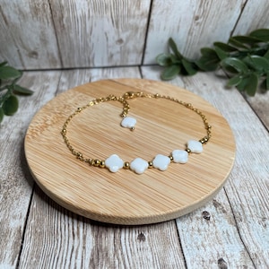 Puede incluir: Un tobillo de cadena dorada con cuentas blancas en forma de trébol. El tobillo está sobre una superficie de madera.