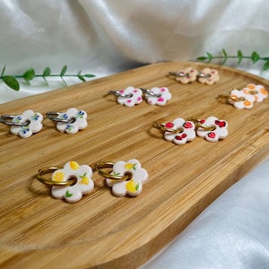May include: A set of 10 flower-shaped earrings with different fruit designs. The earrings are made of white ceramic with gold and silver hoops. The designs include strawberries, lemons, blueberries, and other fruits.