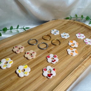 May include: A set of 12 flower-shaped earrings with various fruit designs, including cherries, strawberries, lemons, and oranges. The earrings are displayed on a wooden surface with silver and gold hoop earrings.
