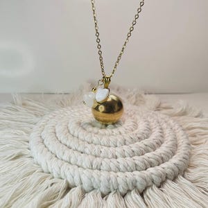 Könnte beinhalten: Eine goldfarbene Halskette mit einem runden Anhänger und einer filigranen Kette. Der Anhänger hat einen kleinen weißen Akzent und hängt an einer goldenen Kette. Die Halskette liegt auf einem weißen, kreisförmigen Makramee-Untersetzer.