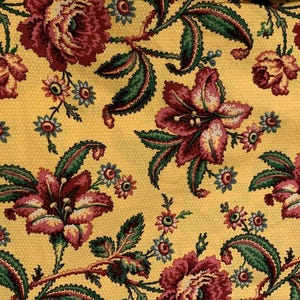 May include: Yellow fabric with a floral pattern. The design features large, burgundy and cream-colored flowers, green leaves, and small blue and cream-colored flowers. The background has a dotted texture.