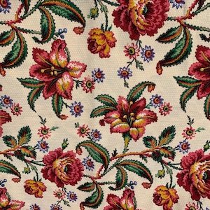 May include: Cream-colored fabric with a repeating floral pattern. The design features large red and yellow flowers, green leaves, and small blue and pink blossoms. The fabric has a textured appearance.
