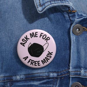 May include: A round, light pink button with the text "ASK ME FOR A FREE MASK" in black. A black KN95 mask graphic is in the center. The button is on a blue denim jacket.