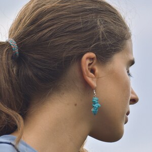 Op de afbeelding: Een vrouw met lang bruin haar dat in een paardenstaart is geknoopt met een blauw en roze elastiekje. Ze draagt een blauw shirt en heeft een turquoise stenen oorbel in haar rechteroor.