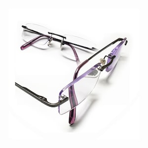 Puede incluir: Un par de gafas sin montura con un marco de metal negro y brazos de plástico morado. Las gafas están dobladas por la mitad y los brazos están cruzados.