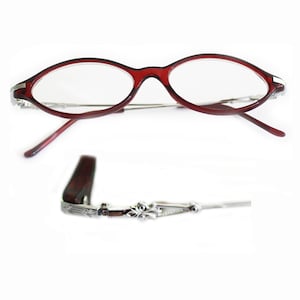 Puede incluir: Gafas de forma ovalada con montura rojo oscuro y patillas plateadas. Las lentes son transparentes y las patillas son finas. Las patillas presentan detalles ornamentales.