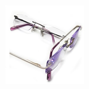 May include: A pair of rimless glasses with a silver frame and purple tinted lenses. The glasses are folded in half and the lenses are facing the camera.