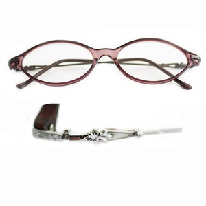 Puede incluir: Un par de gafas ovaladas con montura color burdeos y patillas de metal plateado. Las patillas tienen un diseño floral decorativo.