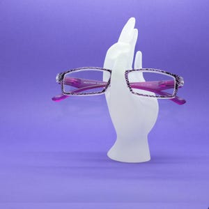 May include: A pair of reading glasses with purple and black patterned frames and bright purple temples. The glasses are held by a white mannequin hand display. The text Montana Eyewear is printed on the inner side of the temple arm.