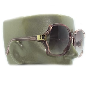 May include: Large, vintage-style sunglasses with a pale pink frame and dark lenses. The sunglasses have a gold-coloured accent on the temples. The glasses are displayed on a green mannequin head, highlighting their shape and design.