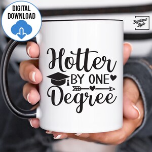 May include: White coffee mug with a black rim and handle. The mug has black text that reads "Hotter by one degree" with a graduation cap, arrow, and hearts.