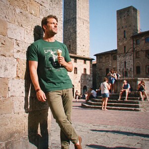 Könnte beinhalten: Ein dunkelgrünes Kurzarm-T-Shirt mit einem weißen, rechteckigen Grafikdruck und dem Text SAN GIMIGNANO, kombiniert mit einer olivgrünen Hose und braunen Slippern. Das T-Shirt hat einen Rundhalsausschnitt und eine zentrierte Illustration.