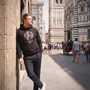 May include: A black hooded sweatshirt featuring a circular white graphic print of a city map and the text FIRENZE. The hoodie has long sleeves, a front kangaroo pocket, and matching drawstrings, worn with dark trousers and white trainers.