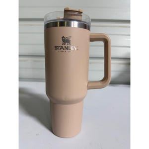 Puede incluir: Un termo de viaje Stanley beige con asa y tapa transparente. El termo presenta el logotipo de Stanley y el texto "SINCE 1913". Está diseñado para mantener las bebidas calientes o frías.