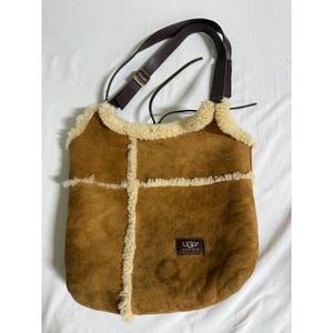 May include: A brown suede hobo bag featuring cream-colored shearling trim along the edges and seams. The bag includes a dark brown adjustable shoulder strap and a small rectangular patch with the text UGG Australia on the lower right side.
