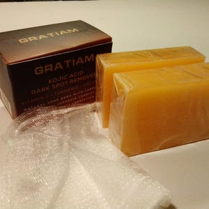 May include: A product box and two bars of soap are displayed. The box is dark brown with the brand name "GRATIAM" in gold lettering. The soap bars are a light orange color and wrapped in clear plastic. A white mesh bag is also present.