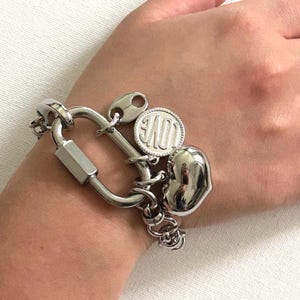 May include: A silver-toned bracelet featuring a large carabiner-style clasp. The bracelet includes charms: a heart, a disc with the word "LOVE", and a small decorative element. The bracelet is designed to be worn on the wrist.