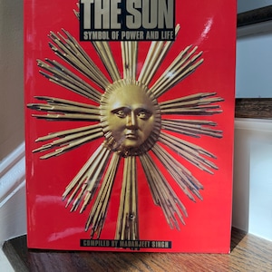 May include: A hardcover book titled "THE SUN" with the subtitle "SYMBOL OF POWER AND LIFE" on a red background. The cover features a golden sun with a face and radiating beams. The author's name, MADANJEET SINGH, is at the bottom.