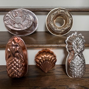 May include: Collection of vintage metal molds for baking, including an eagle, a ring, a pineapple, and a seashell. The molds are in various metallic finishes, including copper and silver, and are displayed on a wooden surface.