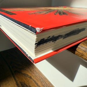 May include: A hardcover book with a bright red cover and a black spine. The pages are white, and the edges show some dark markings. The book is resting on a wooden surface, with a white surface in the background.