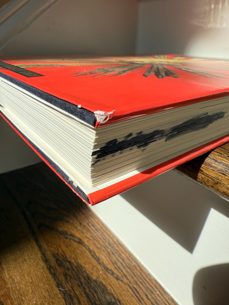 May include: A hardcover book with a bright red cover and a black spine. The pages are white, and the edges show some dark markings. The book is resting on a wooden surface, with a white surface in the background.