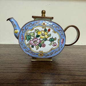 May include: A decorative, round teapot with a blue and white floral design. The teapot features a curved spout, a rounded handle, and a small lid with a gold-colored knob. The floral design includes pink, yellow, and green flowers.
