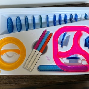 May include: A white plastic drafting tool set with various blue, orange, and pink drawing tools. The set includes a series of blue gears, a pink spirograph, an orange circle template, and several colored pens.
