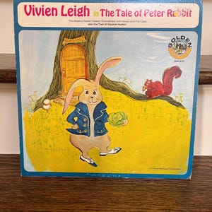 May include: A vintage record album cover featuring a cartoon illustration of Peter Rabbit in a blue jacket holding a green cabbage. The album cover is yellow and green with a blue border. The text on the cover reads "Vivien Leigh in The Tale of Peter Rabbit" and "The Beatrix Potter Classic Dramatized with Music and Full Cast also the Tale of Squirrel Nutkin". The Golden Records logo is in the upper right corner.
