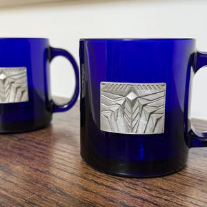 May include: Two cobalt blue glass mugs with silver-colored rectangular embellishments. The embellishments feature a geometric design. The mugs are on a wooden surface.