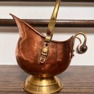 May include: A copper and brass coal scuttle with a decorative handle and a small face on the side. The scuttle is on a wooden surface.