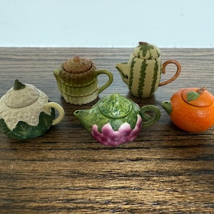 May include: Five miniature teapots in the shape of fruits and vegetables. The teapots include a watermelon, orange, artichoke, and two other teapots with leaf and textured designs. The teapots are made of ceramic and are painted in bright colors.