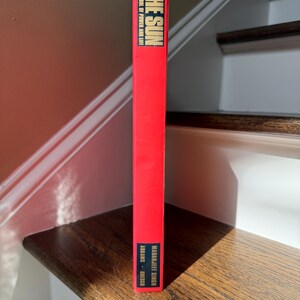 May include: A red book titled "THE SUN" with the text "SYMBOL OF POWER AND LIFE" in white. The author's name, "MADANJIT SINGH", is also visible. The book is standing on a wooden stair step.