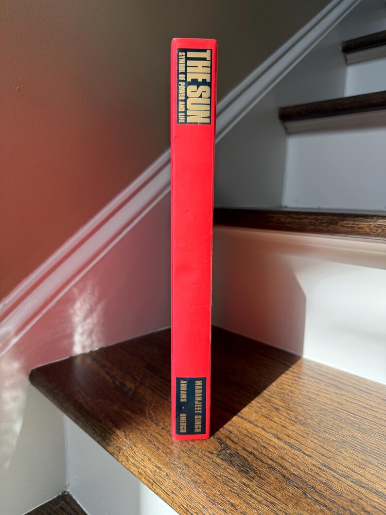 May include: A red book titled "THE SUN" with the text "SYMBOL OF POWER AND LIFE" in white. The author's name, "MADANJIT SINGH", is also visible. The book is standing on a wooden stair step.