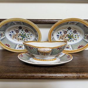 May include: Two oval ceramic bowls and one gravy boat with an attached saucer, all featuring a white base, a yellow and orange border, and a green lattice pattern. The interior and exterior surfaces are decorated with colorful floral motifs.