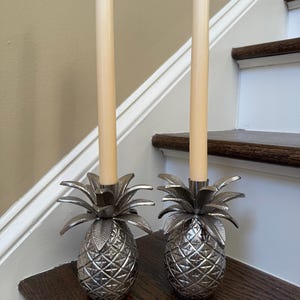 May include: Two silver pineapple-shaped candle holders with cream-colored candles. The candle holders are on a dark wooden surface, with a staircase in the background. The pineapple design is detailed and textured.