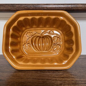May include: A ceramic, pumpkin-shaped baking mold in a warm, earthy brown. The rectangular dish has a scalloped edge and a raised pumpkin design in the center, perfect for fall-themed baking. The mold is approximately 9 inches long.