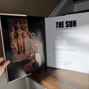 May include: Open book with the title "THE SUN" and the subtitle "SYMBOL OF POWER AND LIFE." The left page features a photograph of a carved stone figure and dancers. The right page contains text and the publisher's information.
