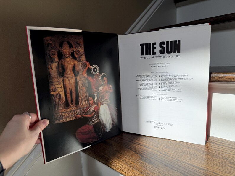 May include: Open book with the title "THE SUN" and the subtitle "SYMBOL OF POWER AND LIFE." The left page features a photograph of a carved stone figure and dancers. The right page contains text and the publisher's information.